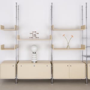 May include: A modular shelving unit with cream cabinets and shelves, supported by silver metal poles. The shelves hold a decorative lamp, a small glass vase with flowers, and a geometric sculpture. The unit is against a white wall.
