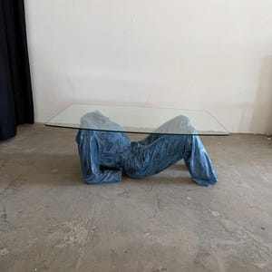 May include: A coffee table featuring a rectangular clear glass top supported by a base shaped like a reclining figure wearing a blue denim jumpsuit. The base is sculpted to resemble the torso and legs of a person lying down in blue fabric.
