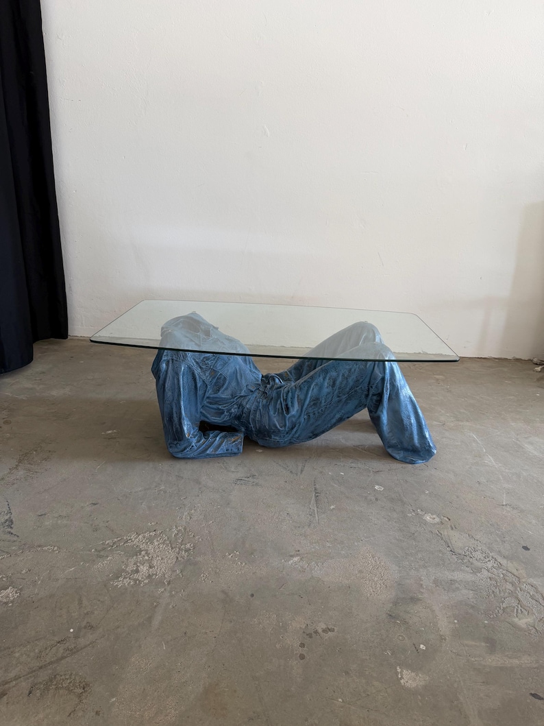 May include: A coffee table featuring a rectangular clear glass top supported by a base shaped like a reclining figure wearing a blue denim jumpsuit. The base is sculpted to resemble the torso and legs of a person lying down in blue fabric.