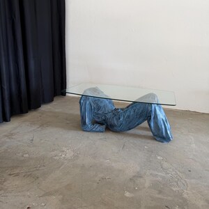 May include: A coffee table featuring a rectangular clear glass top supported by a base shaped like a reclining figure wearing a blue denim jacket and matching blue denim jeans. The base is finished in a metallic blue tone with visible fabric texture.