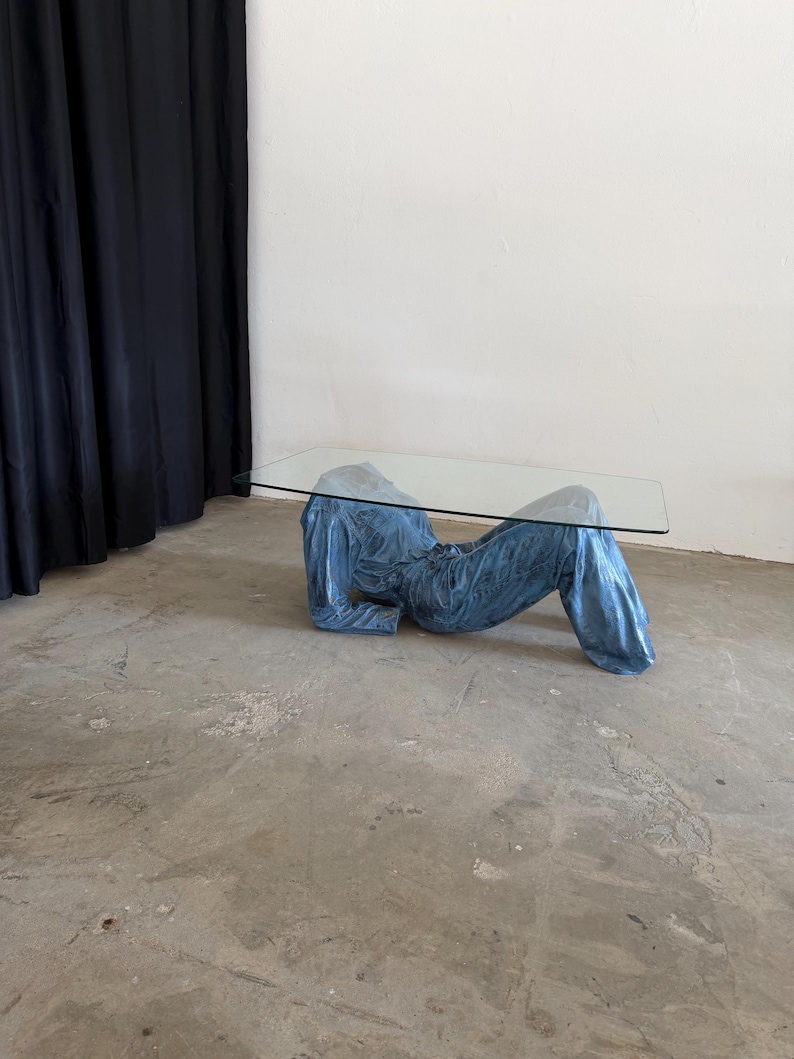 May include: A coffee table featuring a rectangular clear glass top supported by a base shaped like a reclining figure wearing a blue denim jacket and matching blue denim jeans. The base is finished in a metallic blue tone with visible fabric texture.
