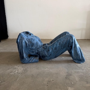 May include: A blue denim jumpsuit sculpture positioned in a reclining pose. The garment features a collared neckline, long sleeves, and a button-down front, with a textured, weathered finish throughout the fabric that mimics a worn appearance.