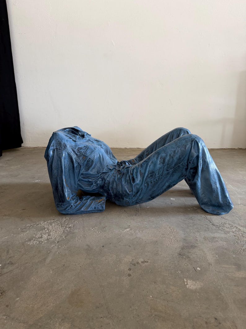 May include: A blue denim jumpsuit sculpture positioned in a reclining pose. The garment features a collared neckline, long sleeves, and a button-down front, with a textured, weathered finish throughout the fabric that mimics a worn appearance.