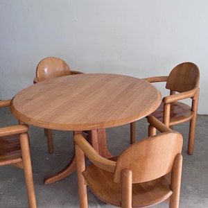 Könnte beinhalten: Ein helles Esszimmer-Set aus Holz mit einem runden Tisch und vier passenden Stühlen. Der Tisch hat einen Sockel, und die Stühle haben geschwungene Rückenlehnen und Armlehnen. Das Set ist aus hellem Holz gefertigt, wahrscheinlich Ahorn oder Birke.