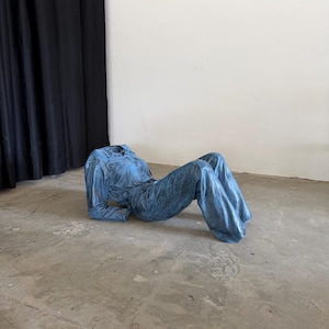 May include: A blue denim jumpsuit with a collared neckline, long sleeves, and wide-leg trousers. The garment is structured to maintain a reclining pose, featuring visible stitching details, a button-down front, and a textured, metallic-like finish.