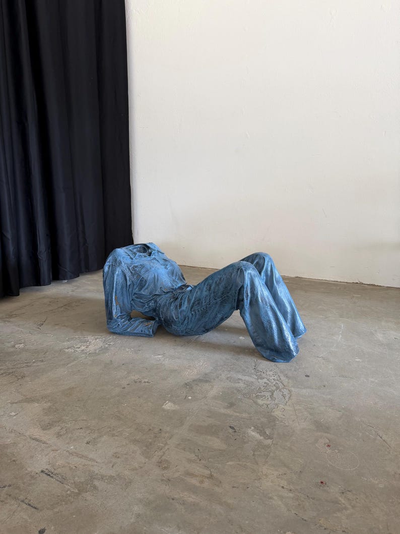 May include: A blue denim jumpsuit with a collared neckline, long sleeves, and wide-leg trousers. The garment is structured to maintain a reclining pose, featuring visible stitching details, a button-down front, and a textured, metallic-like finish.