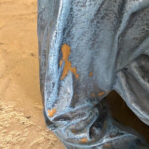 May include: A sculptural object with a textured, metallic blue-grey finish featuring deep folds and creases. The surface shows areas of wear where the blue-grey paint has chipped away to reveal a warm, mustard-yellow base layer underneath.