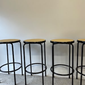 Puede incluir: Cuatro taburetes de metal negro con asientos de caña trenzada. Los taburetes tienen un diseño sencillo con un asiento redondo y un reposapiés en forma de anillo.