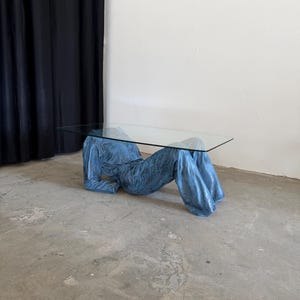 May include: A coffee table featuring a rectangular clear glass top supported by a base shaped like a pair of blue denim jeans and a matching denim jacket, arranged to appear as if a person is lying down while supporting the glass surface.