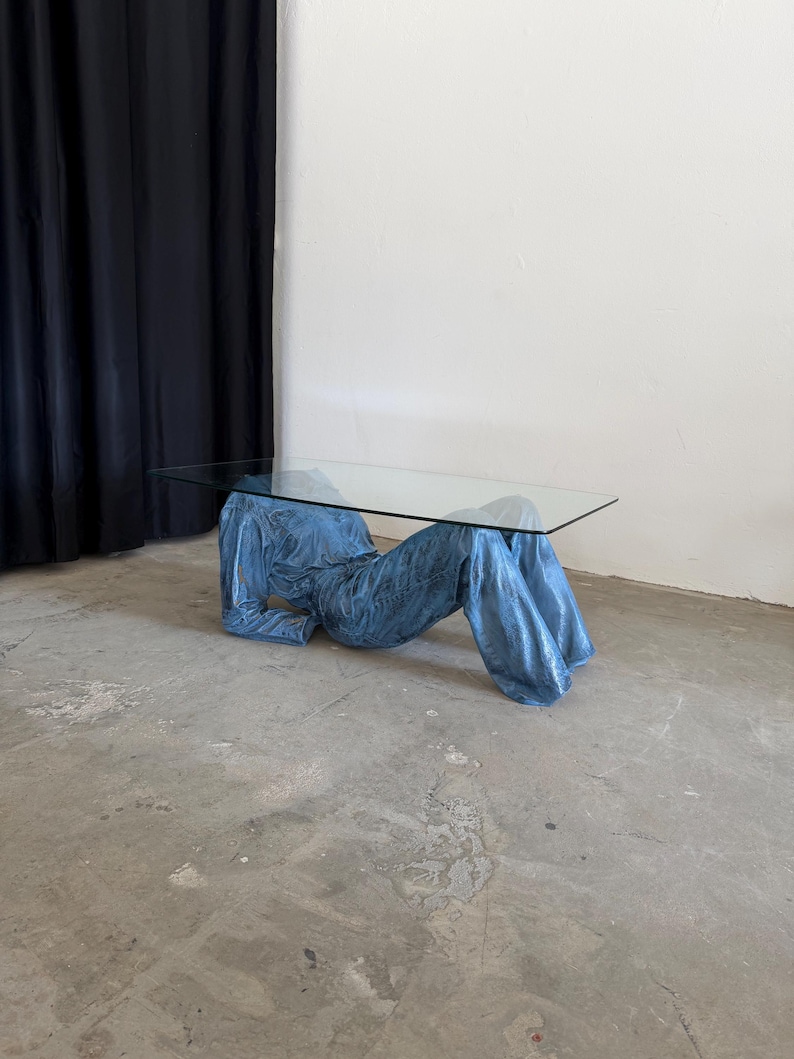 May include: A coffee table featuring a rectangular clear glass top supported by a base shaped like a pair of blue denim jeans and a matching denim jacket, arranged to appear as if a person is lying down while supporting the glass surface.