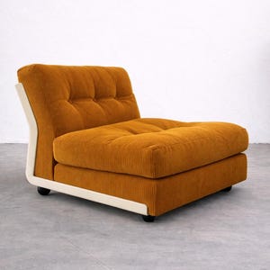 May include: A low-profile armless lounge chair upholstered in mustard yellow corduroy fabric. The chair features a tufted seat and backrest, supported by a curved off-white frame and small black feet, creating a structured and cohesive appearance.