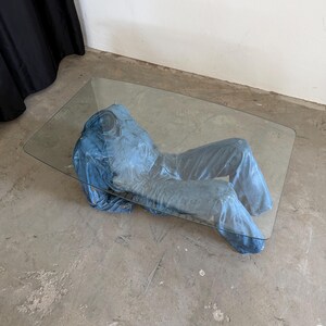 May include: A rectangular coffee table featuring a clear glass top supported by a sculptural base shaped like a blue denim jumpsuit. The jumpsuit appears to be lying flat, with the torso and legs positioned horizontally beneath the glass surface.