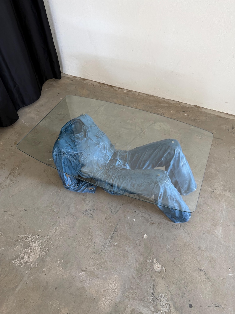 May include: A rectangular coffee table featuring a clear glass top supported by a sculptural base shaped like a blue denim jumpsuit. The jumpsuit appears to be lying flat, with the torso and legs positioned horizontally beneath the glass surface.