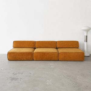 Może przedstawiać: Modułowa sofa składająca się z trzech niskich, prostokątnych sekcji tapicerowanych teksturowaną tkaniną w kolorze musztardowym. Każda sekcja posiada grubą poduszkę siedziska oraz dopasowane oparcie, tworząc ciągły, liniowy układ mebla.