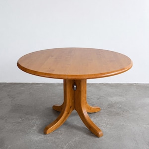 May include: A round wooden dining table with a smooth, light brown finish. The table features a central pedestal base that splits into four curved legs, providing a stable foundation. The tabletop has a visible seam down the centre.