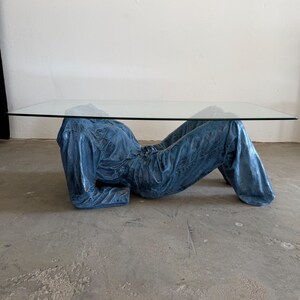 May include: A coffee table featuring a rectangular clear glass top supported by a sculptural base shaped like a reclining figure wearing a blue denim jumpsuit. The base is finished in a textured, metallic blue tone with visible fabric folds.