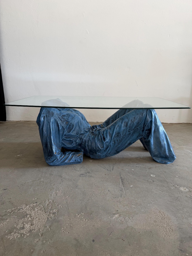 May include: A coffee table featuring a rectangular clear glass top supported by a sculptural base shaped like a reclining figure wearing a blue denim jumpsuit. The base is finished in a textured, metallic blue tone with visible fabric folds.