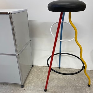 May include: A black leather bar stool with a red, blue, yellow, and white metal frame. The stool has a unique design with a curved yellow metal leg and a circular metal footrest.