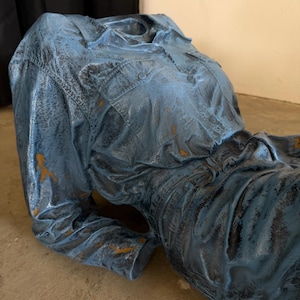 May include: A sculpture of a torso wearing a long-sleeved button-down shirt and pants, finished in a distressed blue paint with dark weathering and patches of exposed brown base material, capturing the texture of fabric folds and seams.