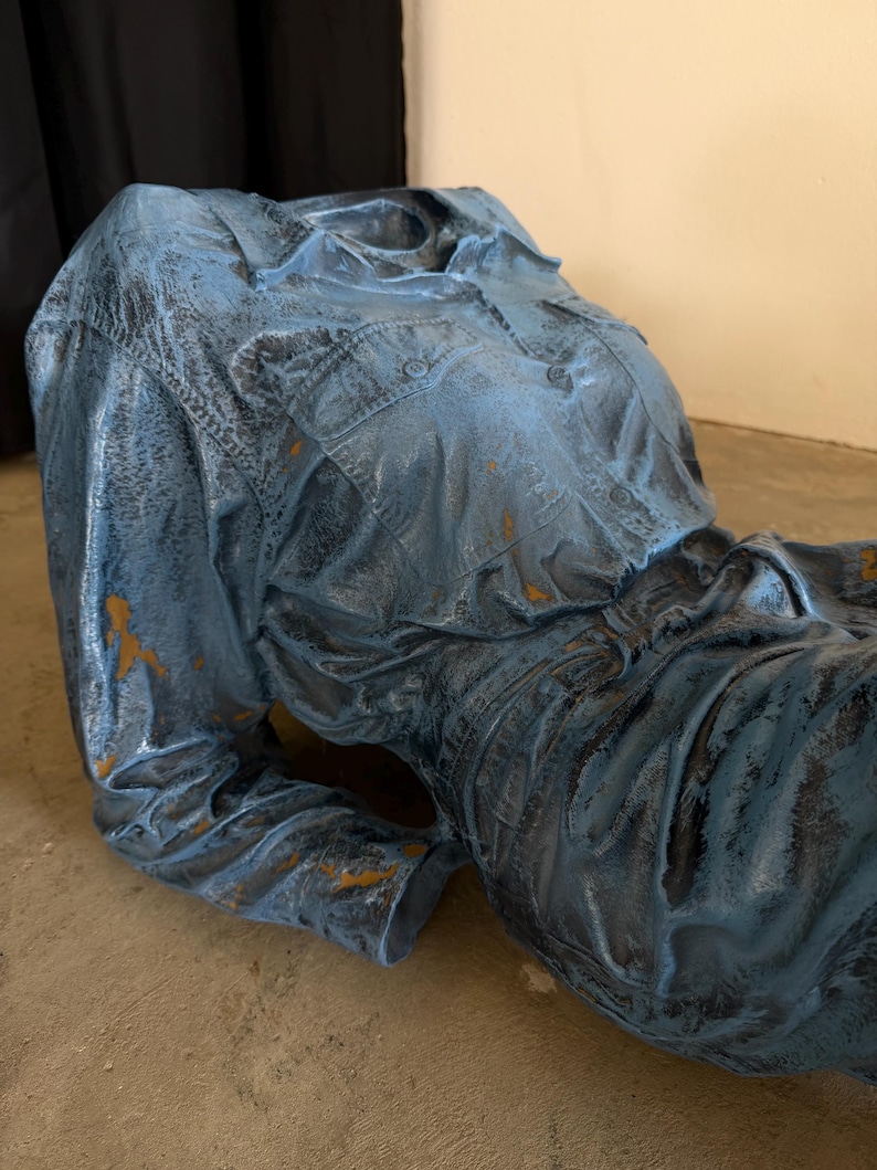 May include: A sculpture of a torso wearing a long-sleeved button-down shirt and pants, finished in a distressed blue paint with dark weathering and patches of exposed brown base material, capturing the texture of fabric folds and seams.