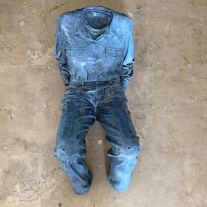 May include: A sculpture of a long-sleeve button-down shirt and trousers in a distressed, weathered blue finish. The garment set features a collared neckline, two chest pockets, and a textured, mottled surface appearance throughout the entire piece.