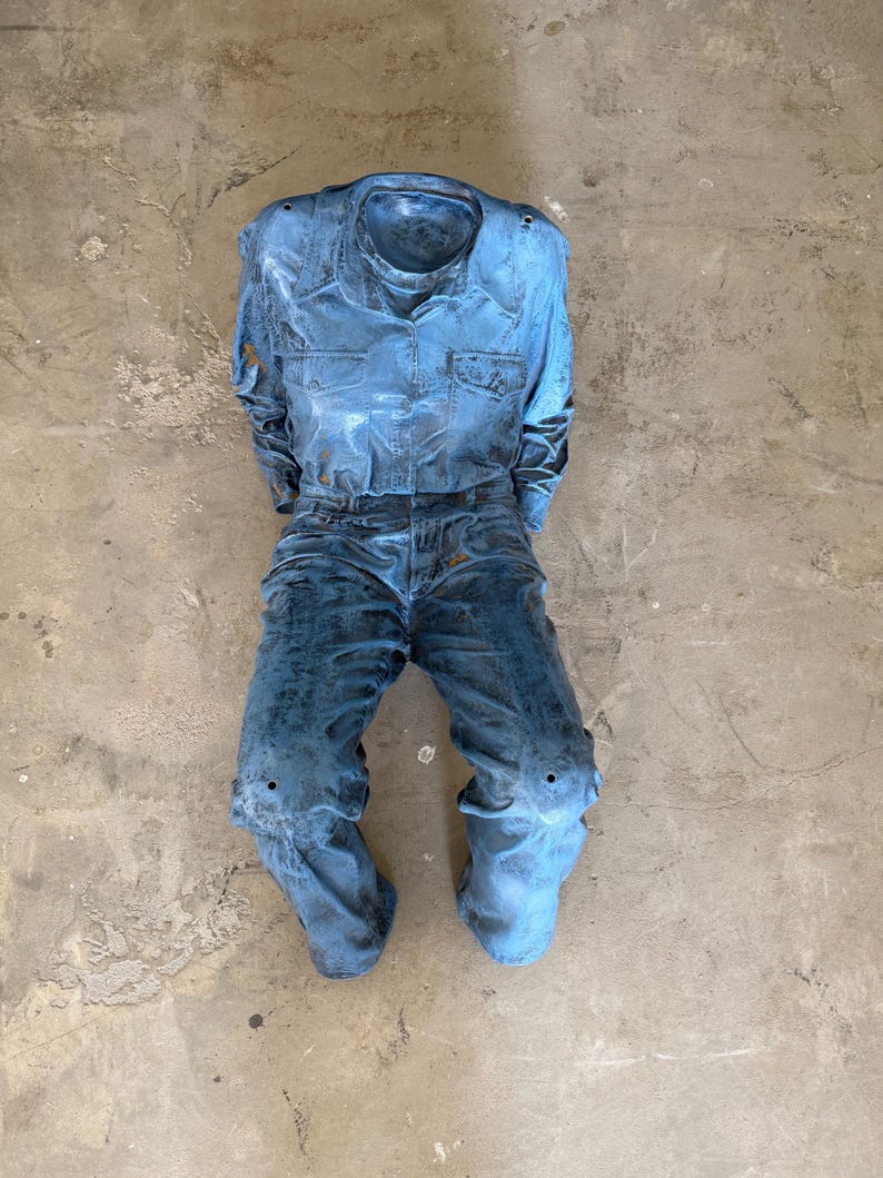 May include: A sculpture of a long-sleeve button-down shirt and trousers in a distressed, weathered blue finish. The garment set features a collared neckline, two chest pockets, and a textured, mottled surface appearance throughout the entire piece.