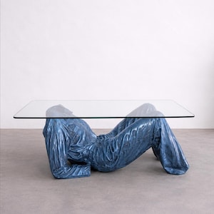 May include: A coffee table featuring a rectangular clear glass top supported by a sculptural base shaped like a reclining figure wearing a blue denim shirt and matching blue denim pants, with a textured, metallic-like finish on the clothing.