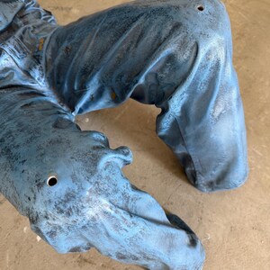 May include: A pair of blue pants with a distressed, weathered finish and a textured appearance. The fabric features visible folds and creases, with a small, circular hole located on each knee area, showcasing a worn and rugged aesthetic.