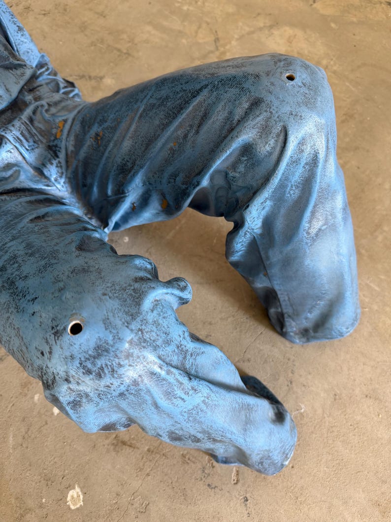 May include: A pair of blue pants with a distressed, weathered finish and a textured appearance. The fabric features visible folds and creases, with a small, circular hole located on each knee area, showcasing a worn and rugged aesthetic.