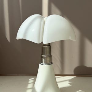 Könnte beinhalten: Eine Tischleuchte mit einem weißen, blütenblattförmigen Glasschirm, der an eine geöffnete Blume erinnert. Die Leuchte verfügt über einen metallischen, abgestuften Mittelschaft und einen glatten, weißen, konischen Fuß, der sich nach unten hin weitet.