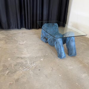 May include: A coffee table featuring a rectangular clear glass top supported by a base shaped like a blue long-sleeved shirt and trousers. The base has a textured, mottled blue finish and is positioned as if a figure is reclining on the floor.