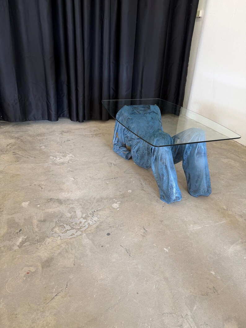 May include: A coffee table featuring a rectangular clear glass top supported by a base shaped like a blue long-sleeved shirt and trousers. The base has a textured, mottled blue finish and is positioned as if a figure is reclining on the floor.