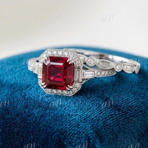 Peut inclure: Une bague en métal argenté avec une pierre précieuse rouge foncé de taille carrée, entourée d'un halo de petits diamants ronds. L'anneau est orné de diamants baguette et ronds, accompagné d'une alliance assortie incrustée de diamants.