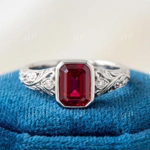 May include: A silver-toned ring featuring a rectangular, emerald-cut red gemstone set in a bezel. The band is crafted with intricate, raised floral and vine-like engravings on both sides, accented by small, round, clear stones embedded within the metal.