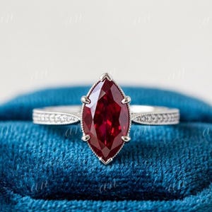 Puede incluir: Un anillo con una piedra preciosa de color rojo intenso en talla marquesa montada en una banda de metal plateado. La piedra central está asegurada por seis garras y la banda presenta un detalle de borde milgrain con pequeñas piedras claras.