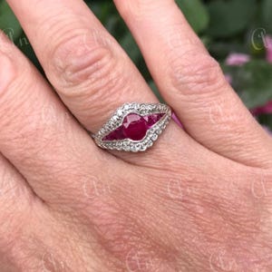 Puede incluir: Un anillo de plata con una gran piedra preciosa de rubí rojo rodeada de diamantes más pequeños. El anillo está engastado en un diseño delicado y curvo.