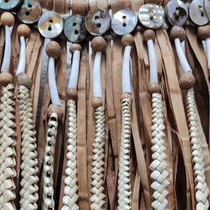 以下が含まれることがあります： 手作りのネックレスのクローズアップ。編み込まれた天然繊維、木製のビーズ、虹色の貝殻ボタンが特徴です。素朴でアースカラーの美しさがあり、さまざまな質感と色合いが混ざり合っています。