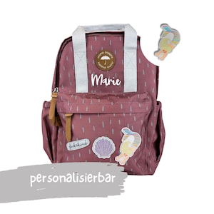 Könnte beinhalten: Ein altrosa Rucksack mit weißen Trägern und dem Namen "Marie" in weißer Schrift. Der Rucksack hat eine Vordertasche, Seitentaschen und dekorative Aufnäher, darunter eine Muschel und eine Cartoonfigur. Der Text "personalisierbar" steht unten.