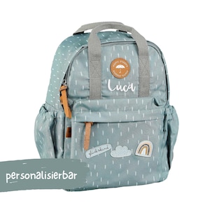 Könnte beinhalten: Ein hellblauer personalisierter Rucksack mit einem weißen Regenmuster und einem weißen Patch, auf dem "glückkind" steht. Der Rucksack hat einen braunen Lederriemen und einen weißen Patch mit dem Namen "Luca".