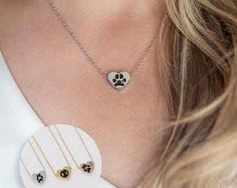 Puede incluir: Un collar de plata con un pequeño colgante de corazón con una huella de pata. El collar se muestra en una modelo con cabello rubio.