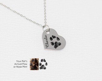 Puede incluir: Collar de plata en forma de corazón con una huella de pata y el nombre "Charlie" grabado en él. El collar está sobre un fondo blanco.