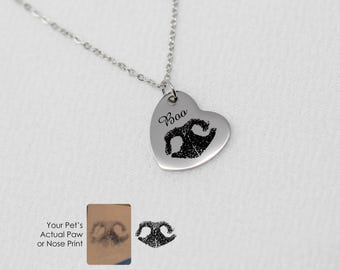 Puede incluir: Collar de plata en forma de corazón con una huella de pata y el nombre "Bao" grabado. El collar se muestra sobre un fondo blanco.