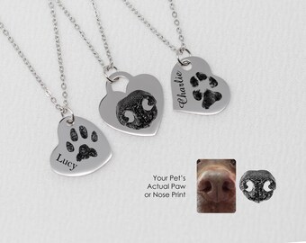 Puede incluir: Tres collares de plata con colgantes en forma de corazón. Cada colgante presenta una huella de pata y un nombre: Lucy, Charlie y un tercer nombre no es visible. El texto "La huella de pata o nariz real de tu mascota" está debajo de los colgantes.