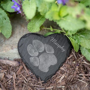 Puede incluir: Una lápida con forma de corazón de pizarra negra para mascotas con una huella de pata y el nombre "Murphy" grabado en letra cursiva. La piedra está rodeada de mantillo y vegetación.