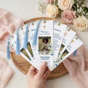 May include: A collection of memorial bookmarks with blue tassels, featuring floral and butterfly designs. Each bookmark includes a cross, the text "Forever in our Hearts", and a photo. The bookmarks are arranged on a wooden tray.