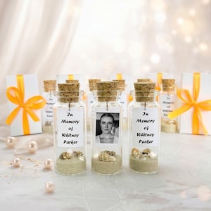 May include: Small glass bottles with cork stoppers, filled with sand and seashells. Each bottle has a label that reads "In Memory of Whitney Parker" and a small photo. White gift boxes with orange ribbons are also present.