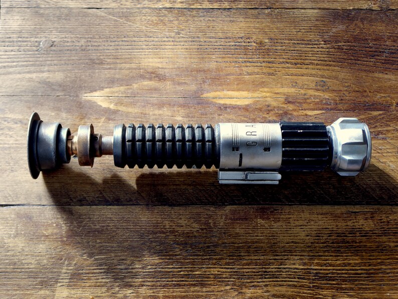 Peut inclure: Un accessoire de sabre laser noir et argent avec une poign&eacute;e noire textur&eacute;e et un canon argent&eacute; avec le mot "GRAH" imprim&eacute; dessus.