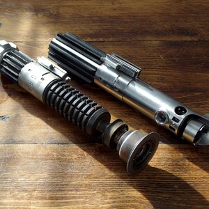 Peut inclure: Un sabre laser argent&eacute; et noir d&eacute;mont&eacute; avec une poign&eacute;e noire et une lame argent&eacute;e. Le sabre laser est sur une surface en bois.
