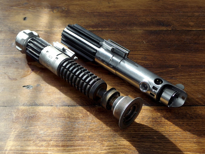 Peut inclure: Un sabre laser argent&eacute; et noir d&eacute;mont&eacute; avec une poign&eacute;e noire et une lame argent&eacute;e. Le sabre laser est sur une surface en bois.