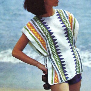 May include: A white, sleeveless tunic with a geometric pattern in blue, green, orange, and yellow. The tunic is paired with purple shorts. The image is taken on a sandy beach with the ocean in the background. The style is vintage.